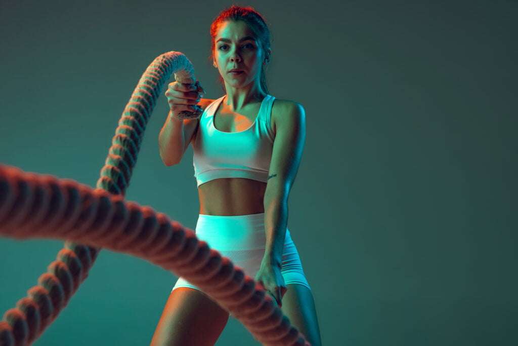 Portrait of young spotive girl doing exercises with rope, keeping body fit isolated over green background in neon
